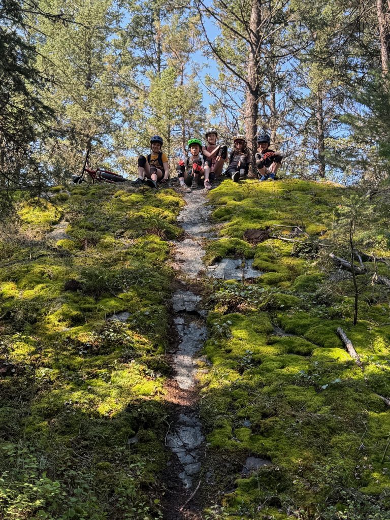 5 kids sitting together on a rock cover in moss with a narrow bike trail going down the middle.